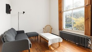 A room with a grey sofa, a wooden chair, a small table with a white tablecloth, a floor lamp, and a window with a view of trees. - New! Bright Brixton 1BR | Walk to Market & Tube (Greater London)