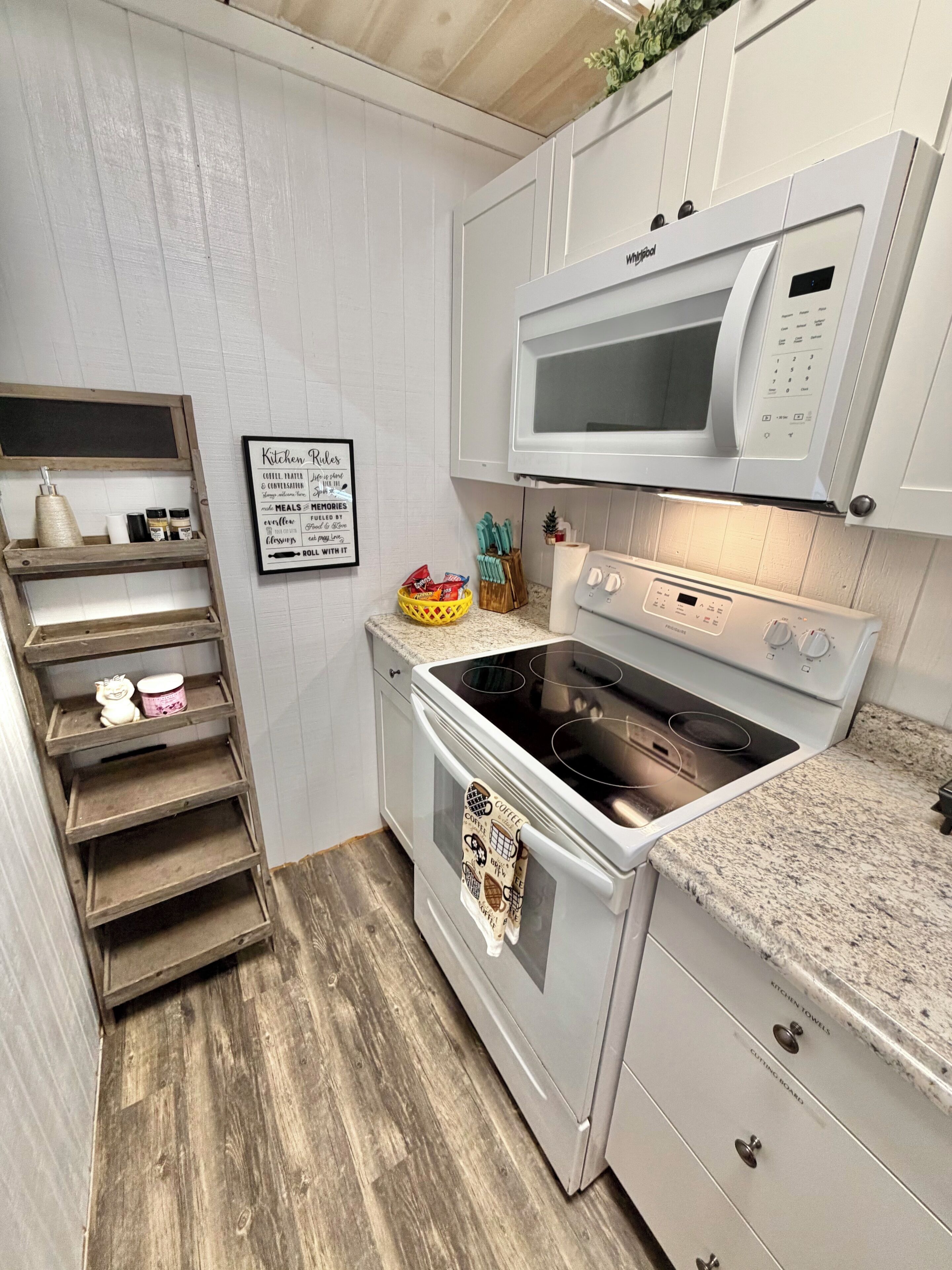 A compact kitchen with a microwave, oven, and a wooden shelf unit.