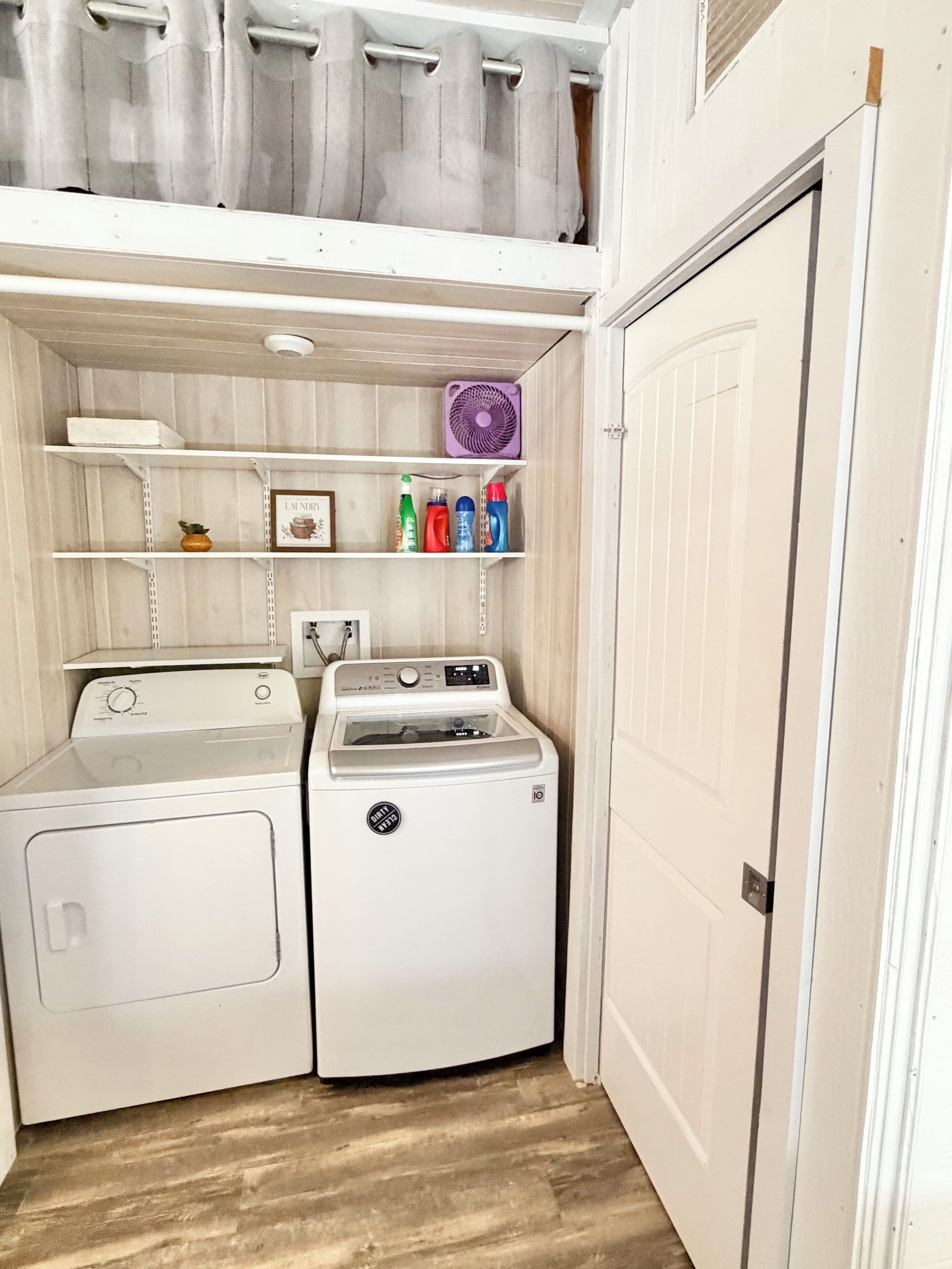 A laundry room with a washing machine, dryer, shelves with cleaning supplies, and a fan.