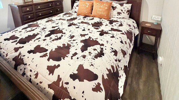 Quarto com cama, cabeceira de madeira, mesa de cabeceira, abajur, um quadro emoldurado e uma obra de arte na parede.