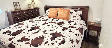 A bedroom with a bed, wooden headboard, a nightstand, a lamp, a framed picture, and a wall-mounted artwork.