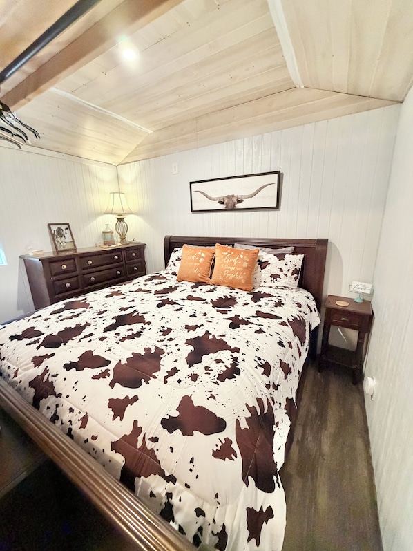 Een slaapkamer met een bed, houten hoofdeinde, nachtkastje, lamp, een lijst met foto en een kunstwerk aan de muur.