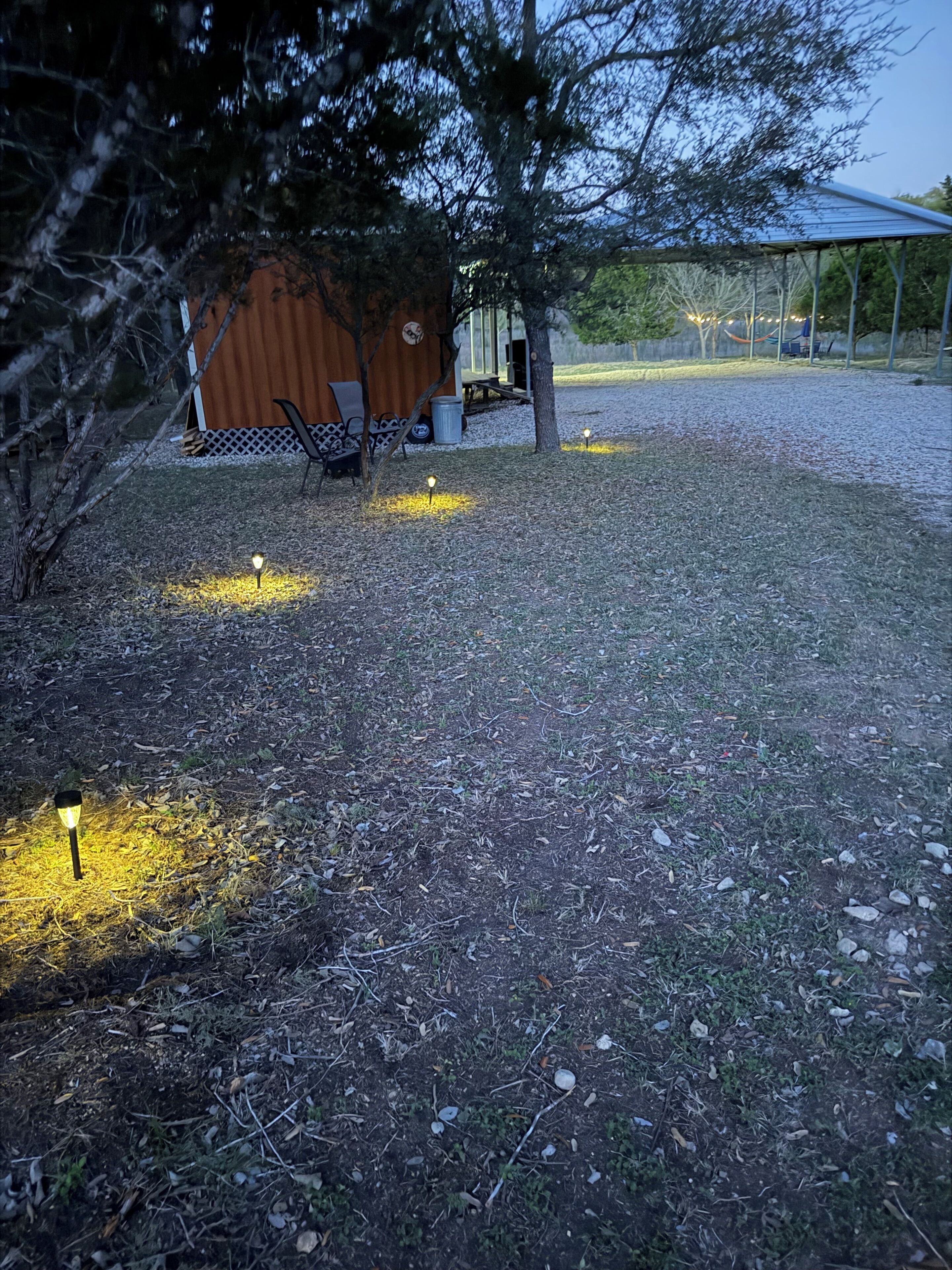 Um quintal com um galpão de madeira, uma área de estar coberta e um gramado com luzes espalhadas.