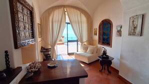 A room with a wooden table, a white sofa, a small side table, and a view through a window with curtains. - Villa Les Paons - Downtown Positano - Terrace (Positano)