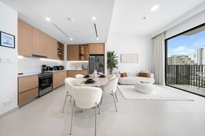 A modern kitchen with built-in appliances, a dining area with white chairs, and a large window with a city view. - StayShort - Dubai Hills - Premium 2BR with Sunlit Balcony - Sleeps 4 (Dubai Hills Estate)