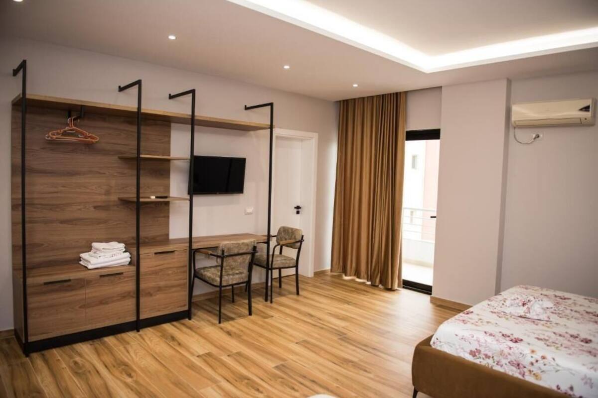 A hotel room with a wooden floor, a bed with a floral bedspread, a TV mounted on the wall, a wooden wardrobe with a hanger, and a table with chairs.