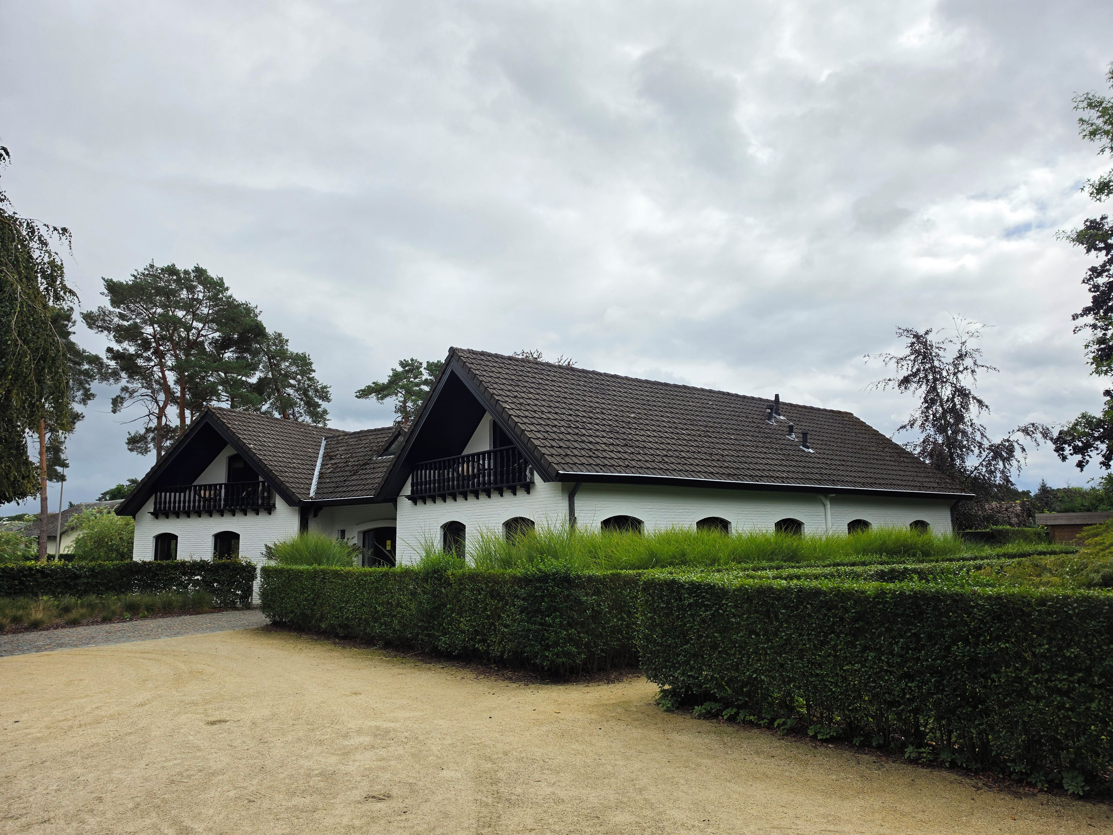 A white building with a tiled roof and multiple windows, surrounded by a well-maintained hedge and a gravel path.