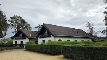 A white building with a tiled roof and multiple windows, surrounded by a well-maintained hedge and a gravel path.
