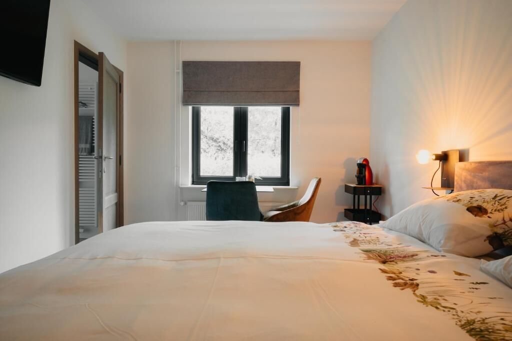 Un dormitorio con cama, una silla, un ventanal, una mesita de noche con lámpara y un televisor montado en la pared.