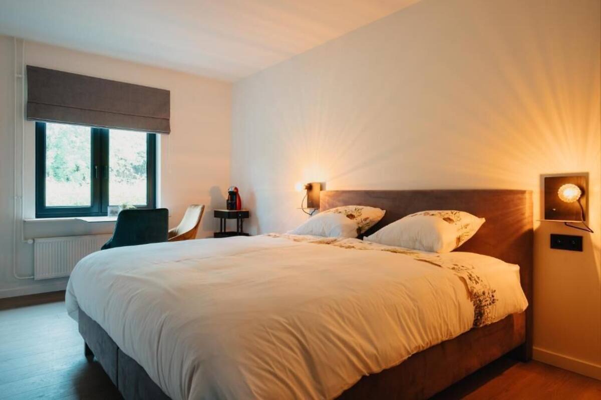 A bedroom with a large bed, a chair, a nightstand, a lamp, and a window with a view of greenery.
