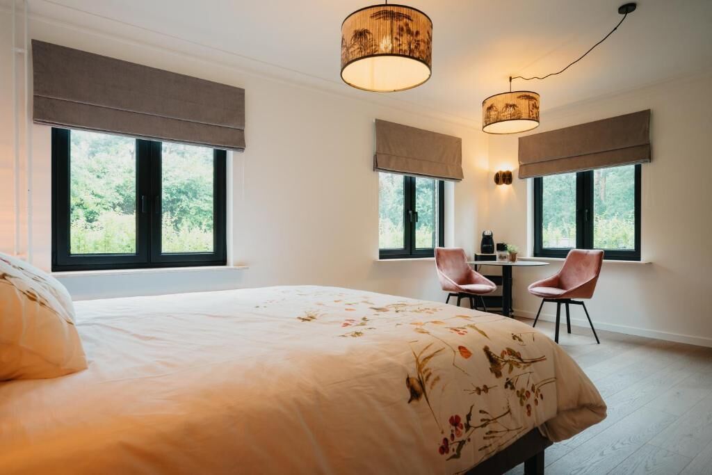 Une chambre à coucher avec un grand lit, deux fauteuils, un bureau et deux fenêtres équipées de stores.