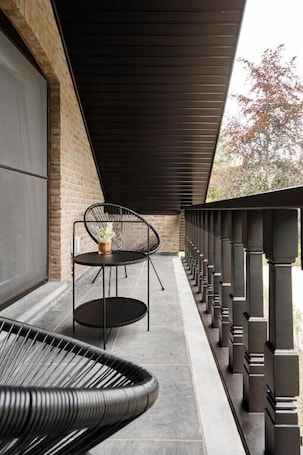 A covered outdoor seating area with a brick wall, a black metal chair, a small round table with a plant, and a black woven chair.. Wilgenhuis