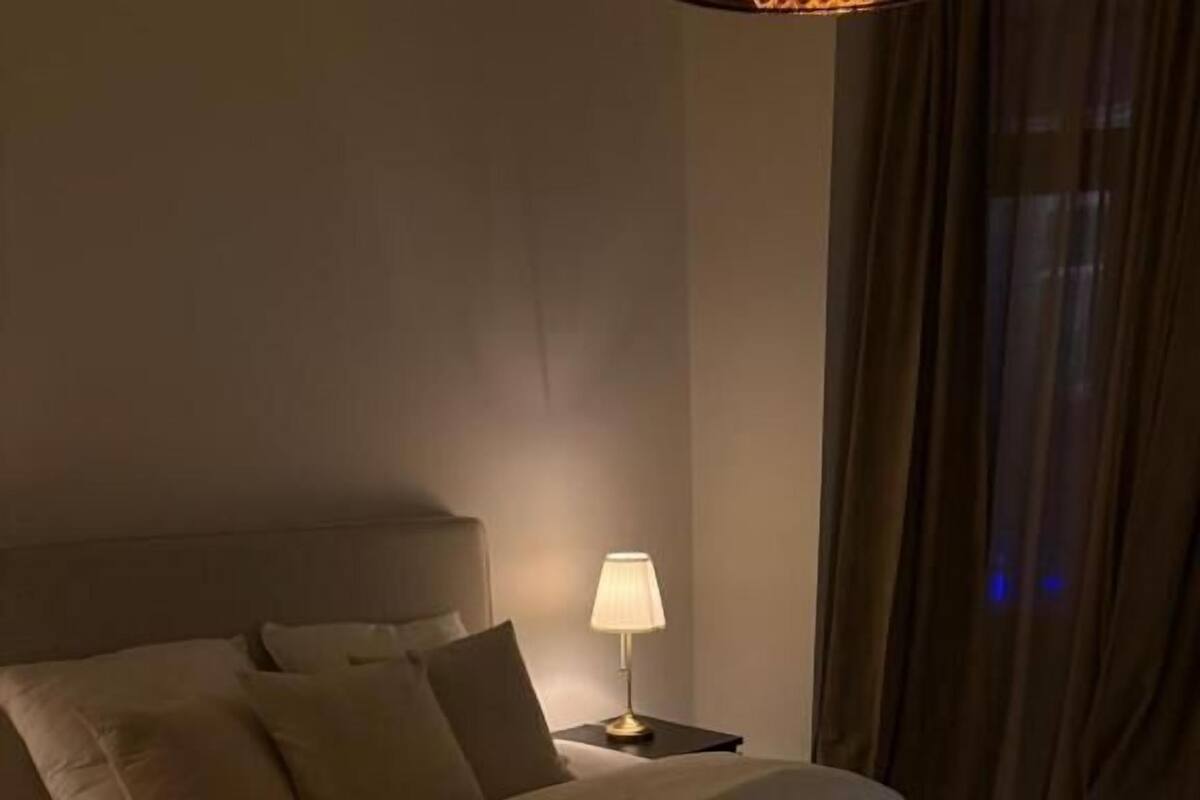 A hotel room with a bed, pillows, a nightstand with a lamp, and a window with curtains.