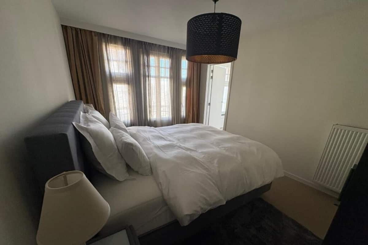 A bedroom with a bed, a bedside lamp, a window with curtains, and a radiator.