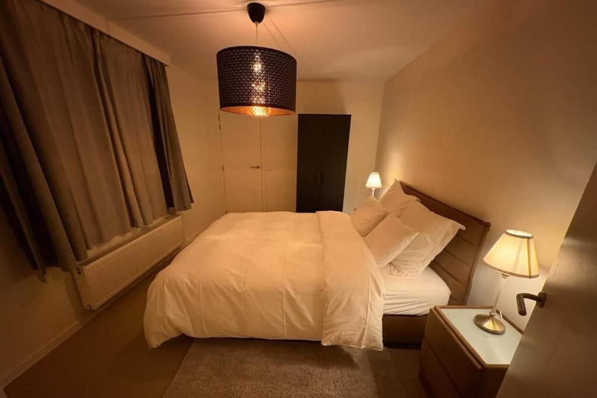 A hotel room with a bed, a nightstand, a lamp, a window with curtains, and a door.
