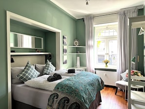 Ein Schlafzimmer mit Bett, Schminktisch, Spiegel, Fenster mit Vorhängen und Heizkörper.