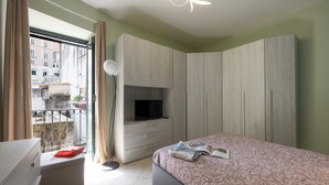 Camera da letto con un letto, una TV su un mobile, un armadio, un balcone con vista sugli edifici e una finestra con tende.