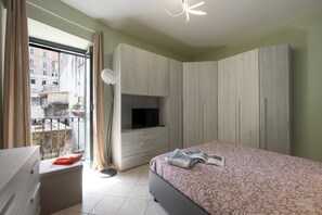 Ein Schlafzimmer mit Bett, Fernseher auf einem Schrank, Kleiderschrank, Balkon mit Blick auf Gebäude und einem Vorhangfenster.
