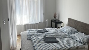 A bedroom with a bed, a bedside table, a lamp, a radiator, and a window with sheer curtains.