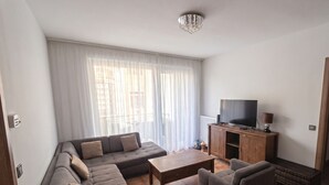 A living room with a sectional sofa, a small table, a TV on a wooden cabinet, and a large window with sheer curtains.