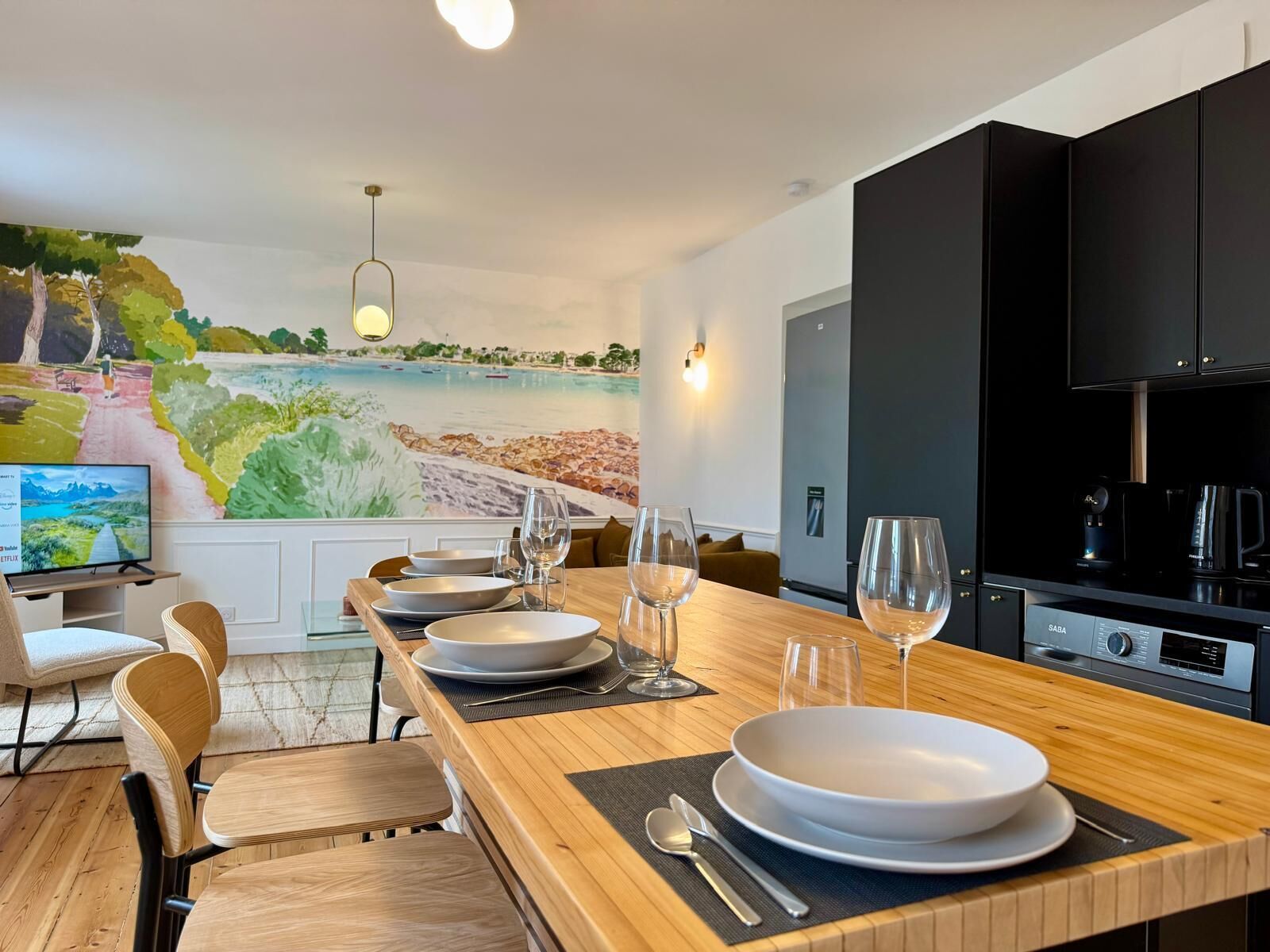 Une cuisine moderne avec un coin repas, un téléviseur et une fresque représentant un paysage côtier.