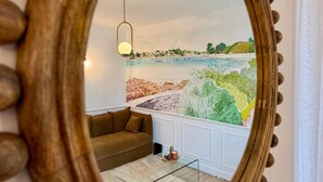 Habitación con un espejo grande que refleja una escena de playa, un sofá marrón, una mesa de centro de vidrio y una lámpara colgante.