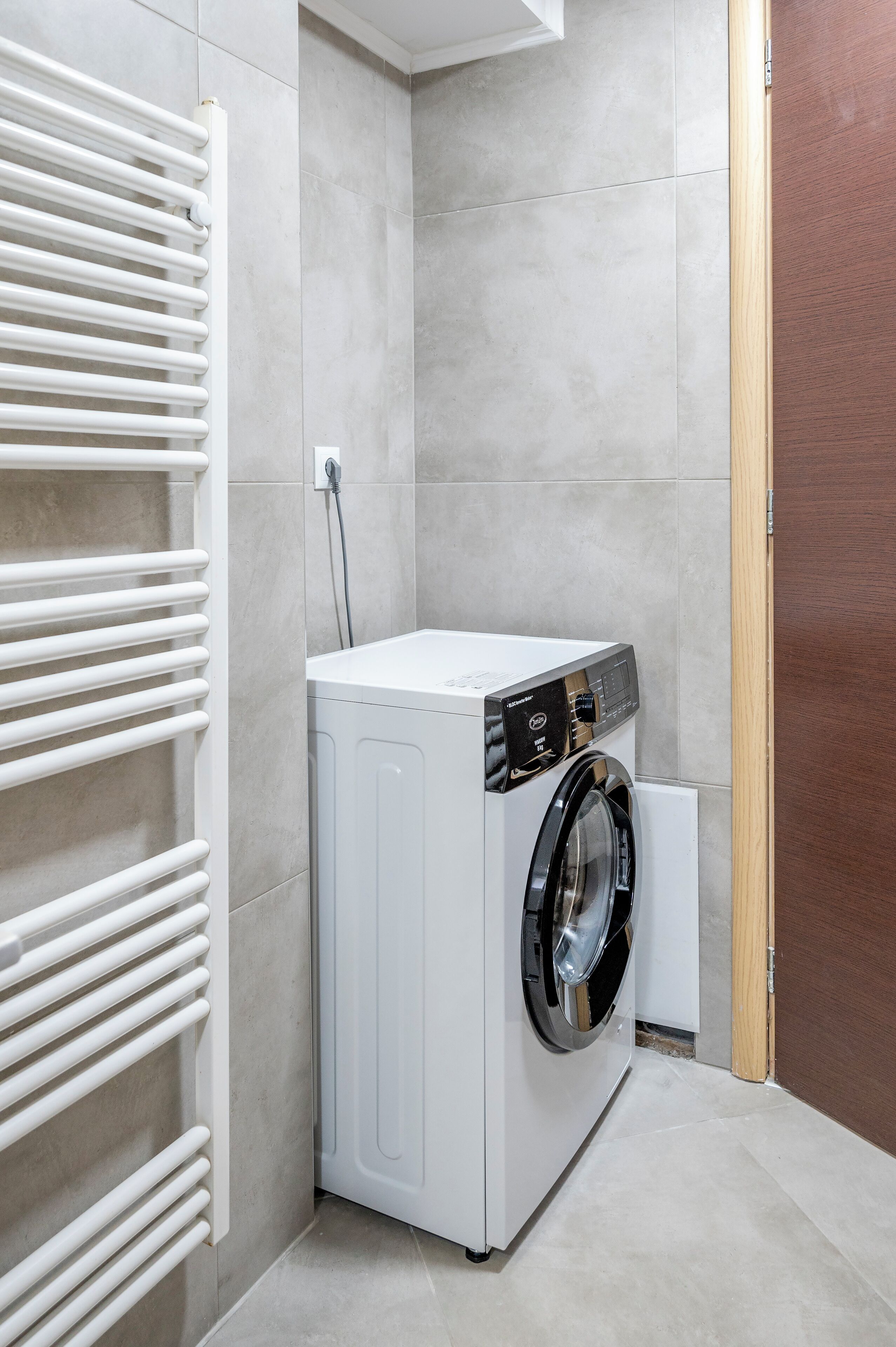 A compact laundry room with a front-loading washing machine, a white radiator, and tiled walls.