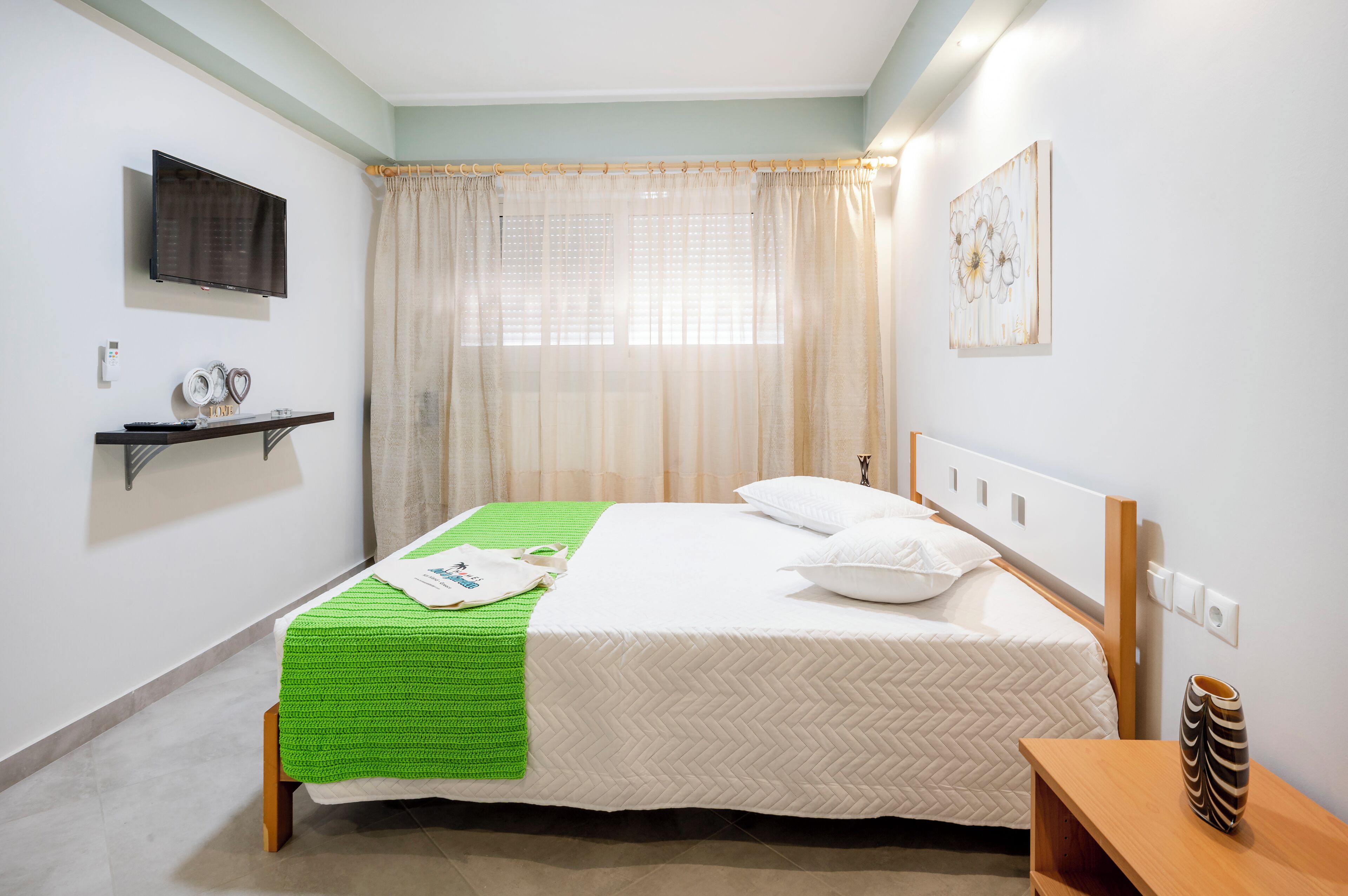Habitación de hotel impecable con cama, mesita de noche, televisor de pared y ventana con cortinas.