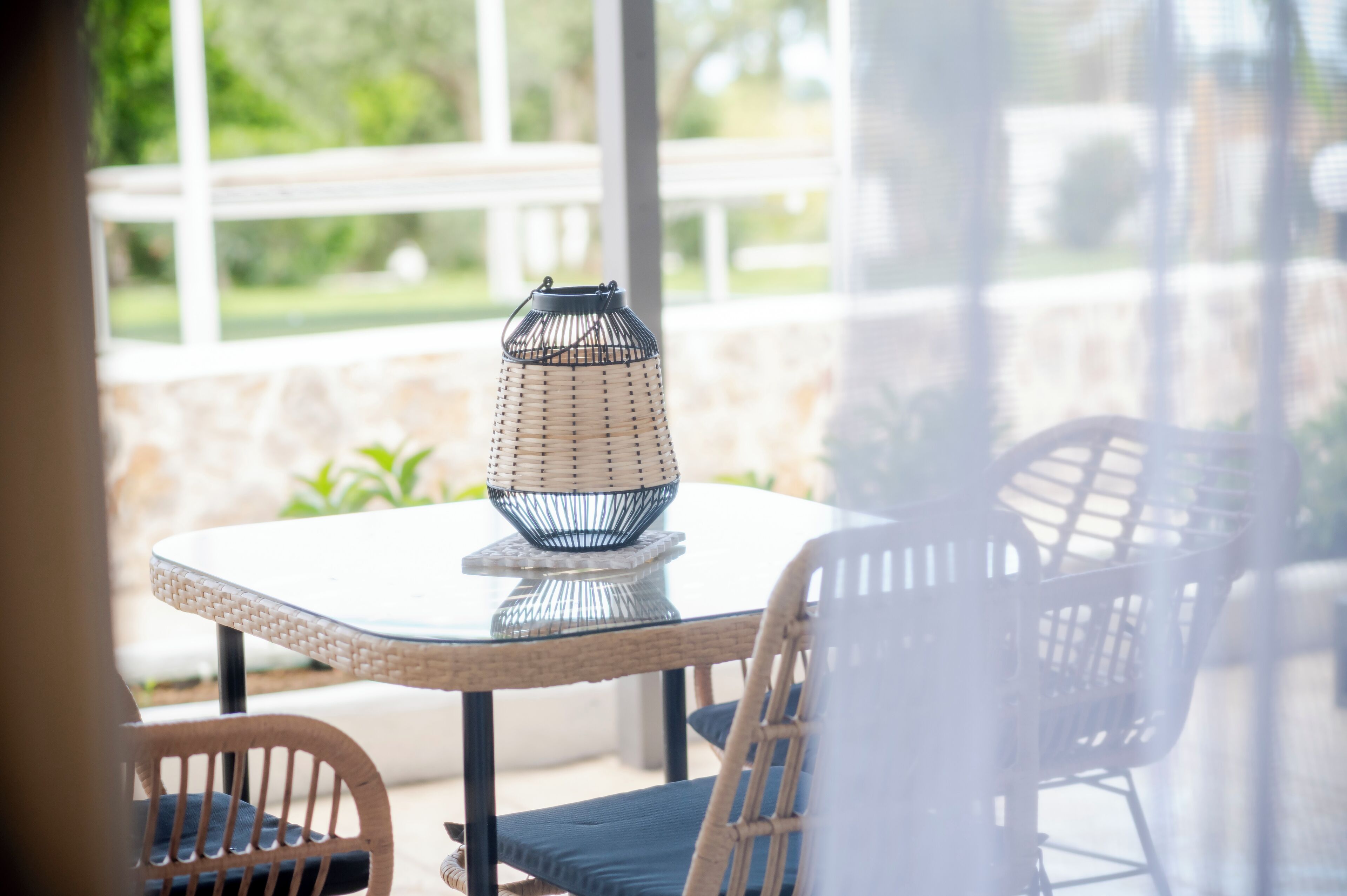Une table dont le plateau est en verre, agrémentée d’un centre de table en forme de panier tressé, de chaises en osier et offrant une vue sur la verdure grâce à de grandes fenêtres.
