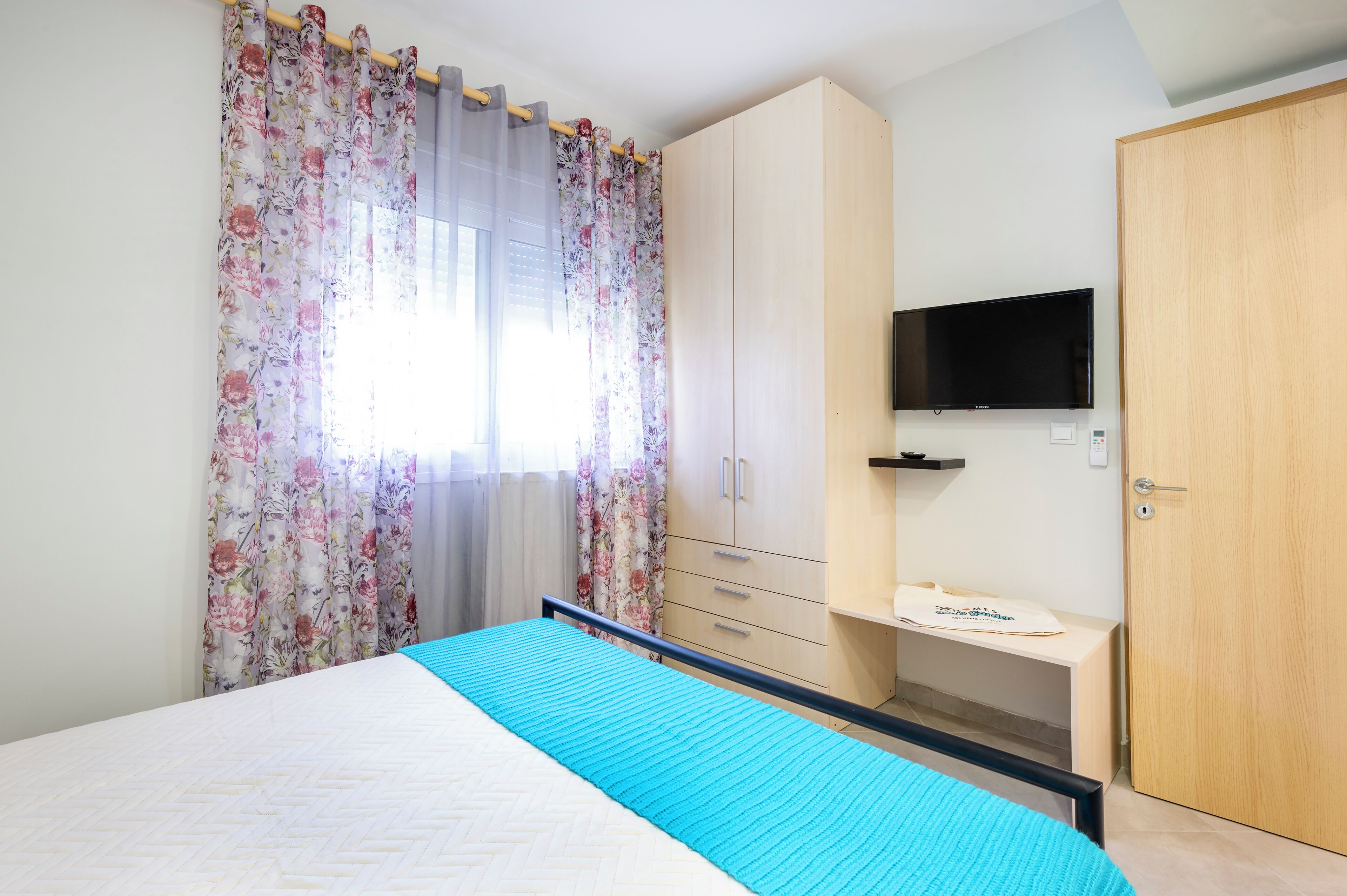 A bedroom with a bed, a TV mounted on the wall, a wardrobe, and a desk.