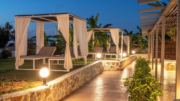 A nighttime outdoor lounge area with cushioned seating under pergolas, stone walls, and a pathway. - Homes Eva's Garden (Kos)