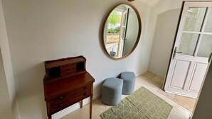 Une pièce comprenant une commode en bois, un miroir rond, deux poufs et une porte donnant sur l’extérieur.