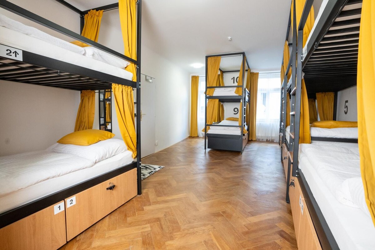 Een slaapzaal met stapelbedden, houten vloeren en gele gordijnen.