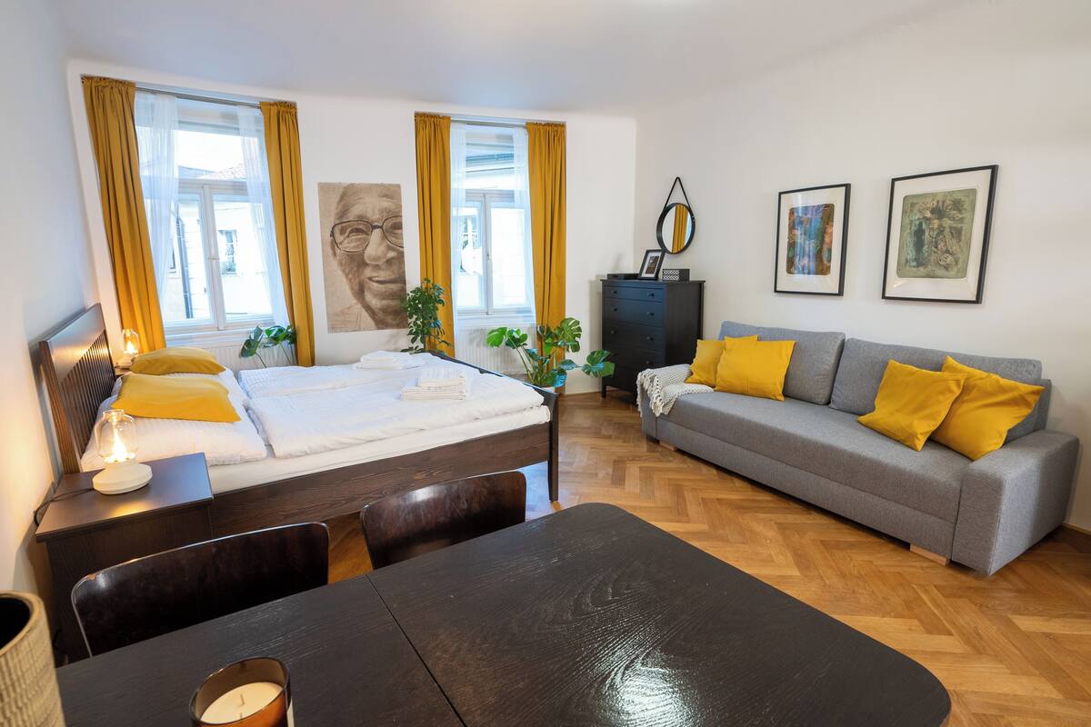 Een moderne slaapkamer met een bed, nachtkastje en een grijze bank met gele kussens.