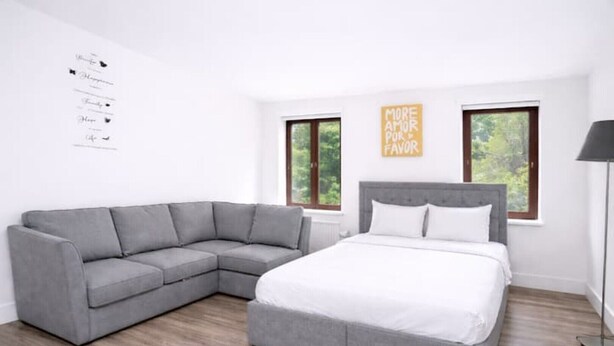 A modern bedroom with a gray sectional sofa, a large bed, two windows, and a wall-mounted sign. - Elegant Flat | 2-Min Walk to Watford Junction (Watford)