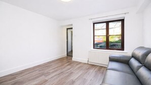 A room with wooden flooring, a black leather sofa, a window with a view, and a door leading to another room. - Elegant Flat | 2-Min Walk to Watford Junction (Watford)
