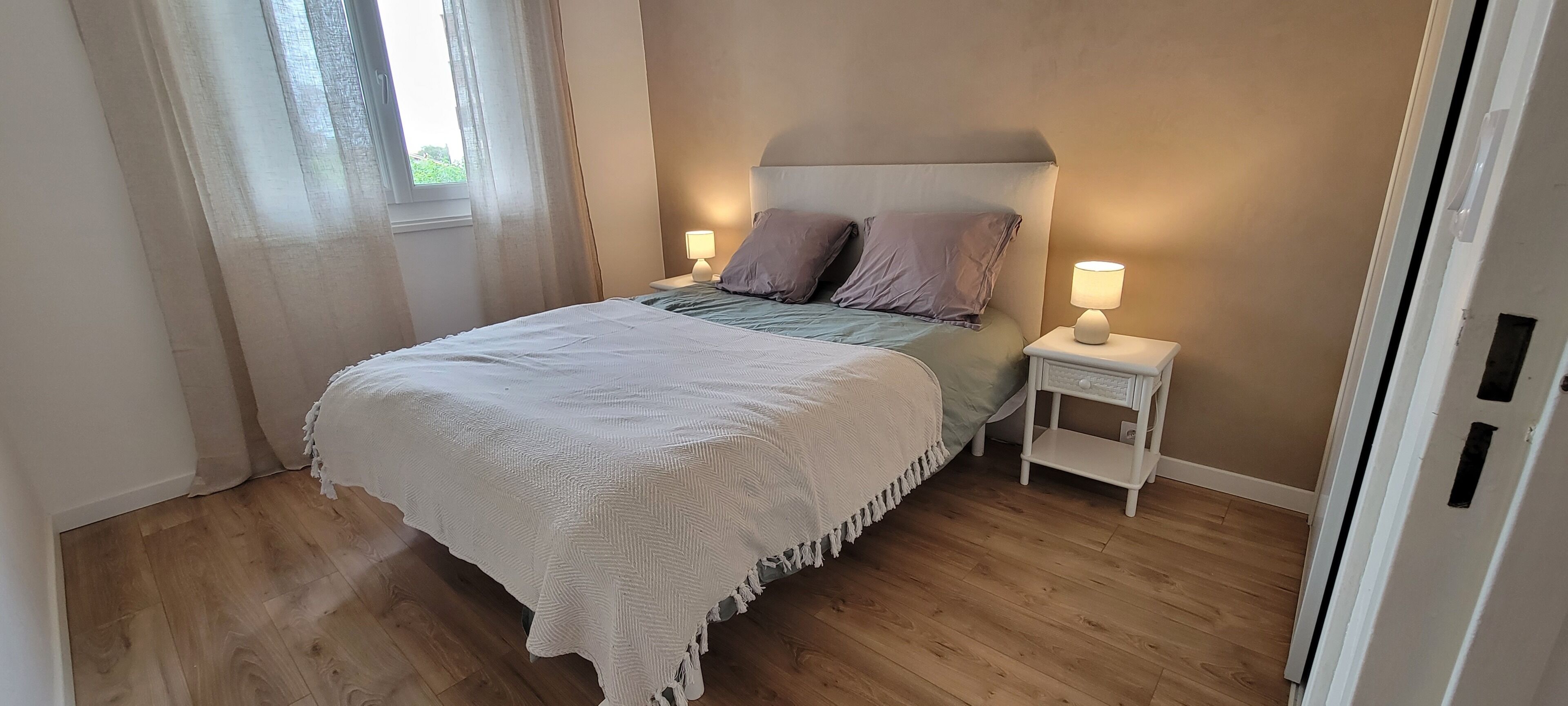 A bedroom with a bed, two bedside tables with lamps, a window with curtains, and wooden flooring.