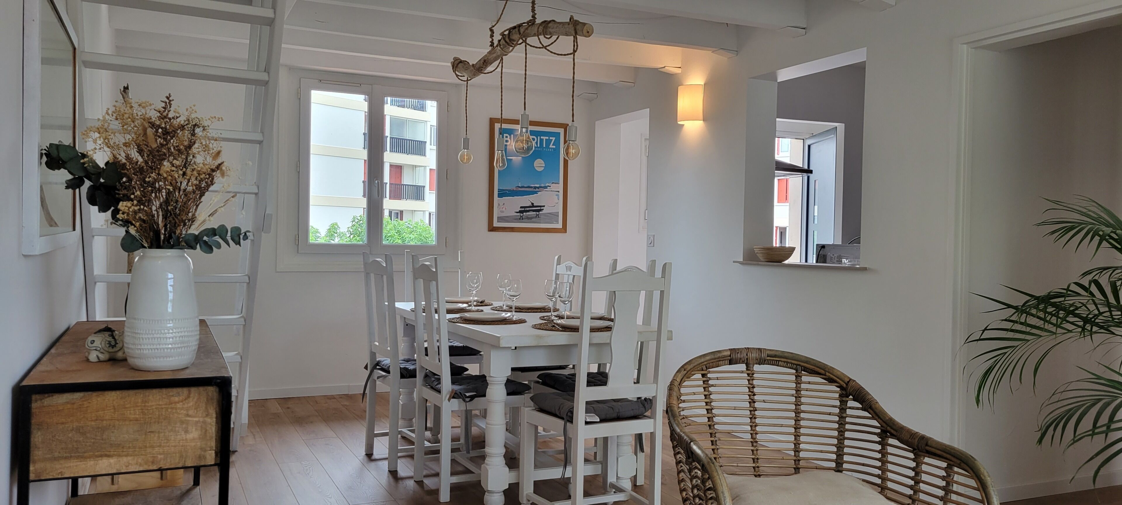 A dining area with a white table set for four, surrounded by white chairs. A wicker chair is positioned to the side. A large white vase with dried flowers is on a side table.