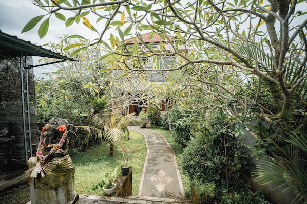 A garden pathway leading to a building with a red roof, surrounded by lush greenery and a statue. - Casa De Arya (Gianyar)