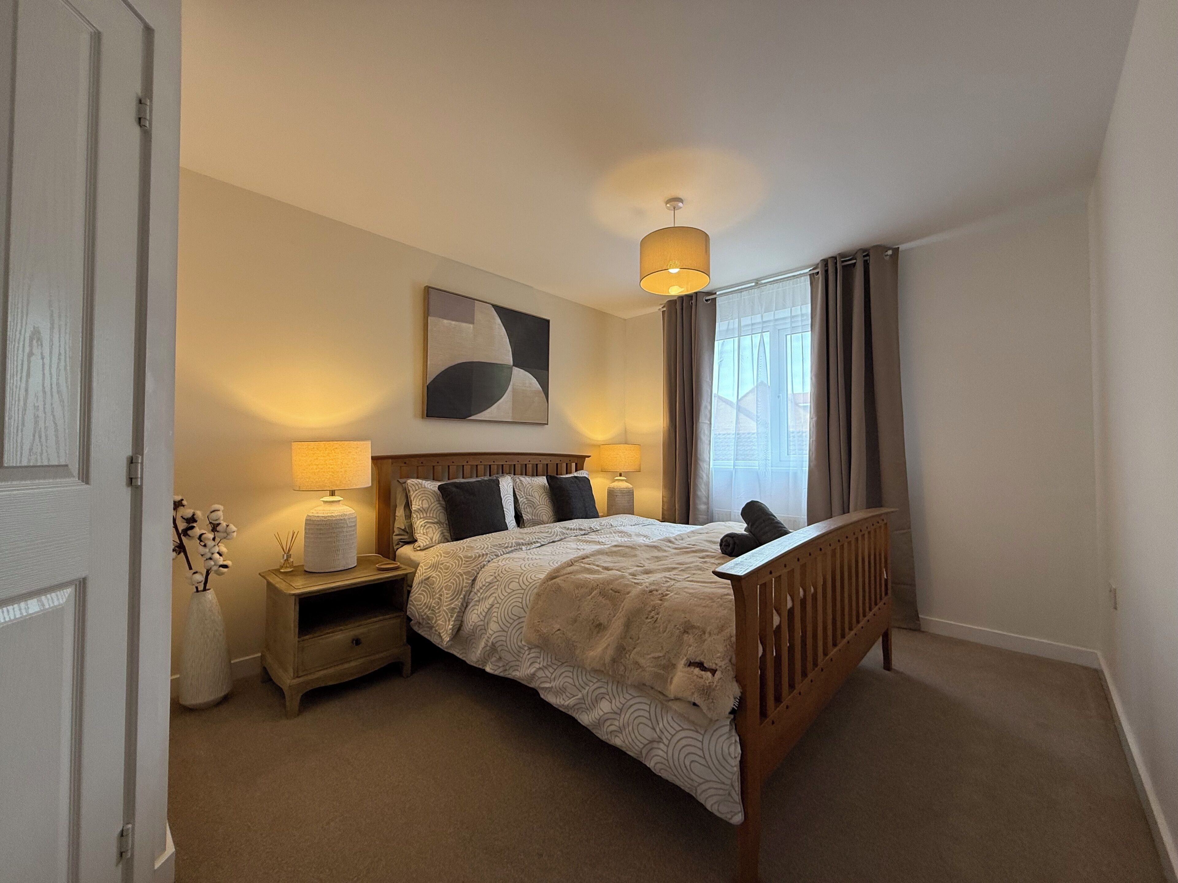 A bedroom with a wooden bed, two bedside tables with lamps, a wall-mounted abstract painting, and a window with curtains.