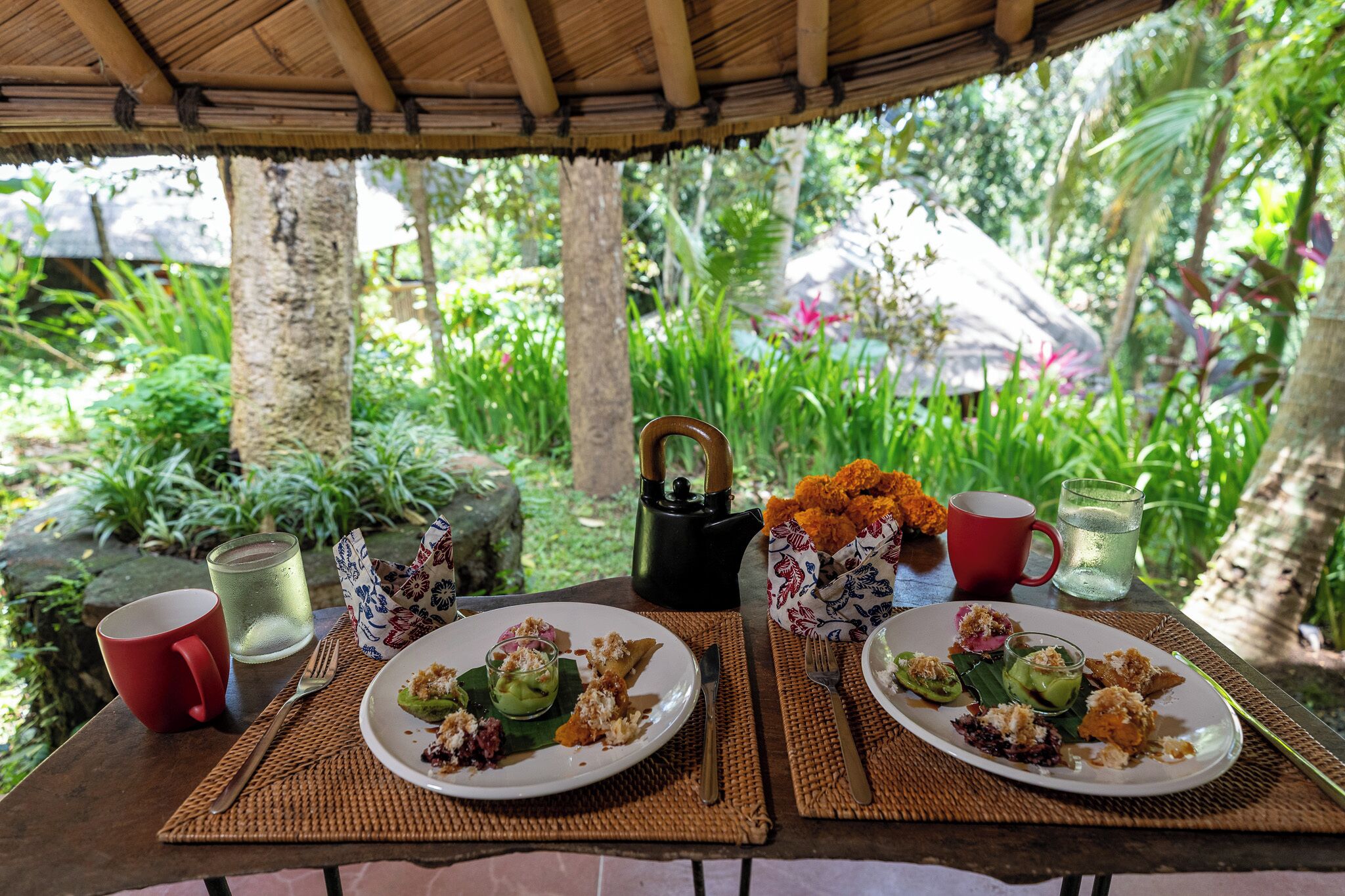 An outdoor dining area with two plates of food, a teapot, a cup, and a glass of water on a table.