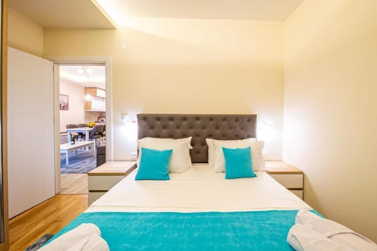 Un dormitorio con una cama grande, dos mesitas de noche, un cabecero capitonado y vistas al salón contiguo.