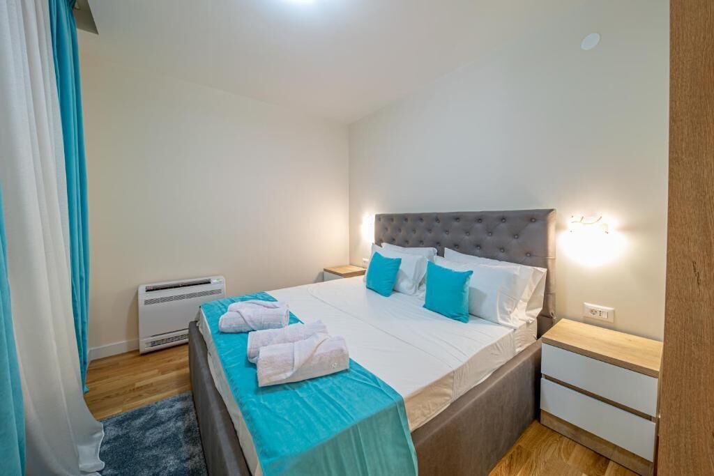 A neatly made bed with a grey headboard, white linens, and blue pillows. There is a bedside table with a lamp, a wall-mounted air conditioner, and a small wooden nightstand.
