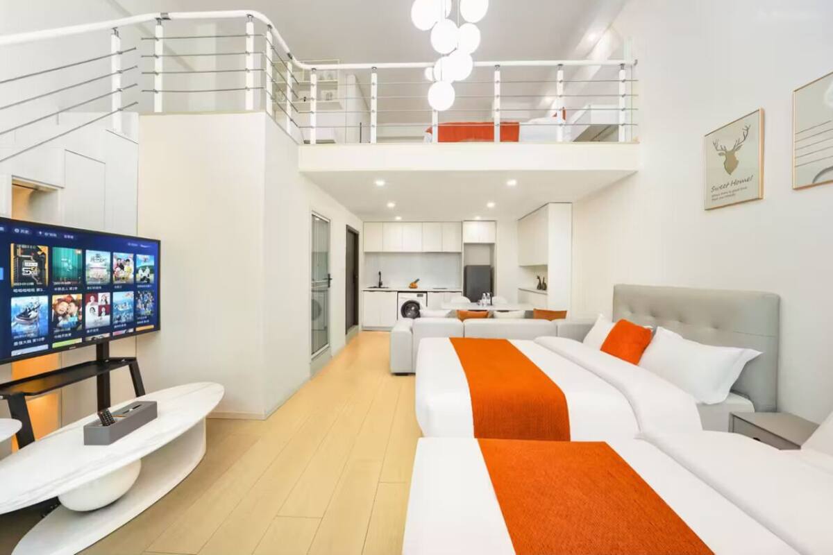 Une chambre d’hôtel moderne dotée d’une télévision à écran plat, d’un lit avec une literie orange, d’un canapé, d’un coin repas et d’une cuisine.