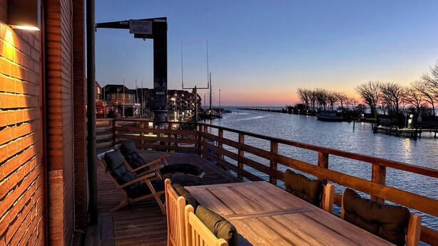 A wooden deck with outdoor seating, a view of the water, and a building with a sign. - Pirate ship-private spa, fireplace, sea view & gym (Fehmarn)