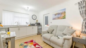 A modern living room with a sofa, a dining table, and a kitchen area with a sink and a clock. - Apartment Sleeps 2 With Parking and Pets Welcome (Narberth)