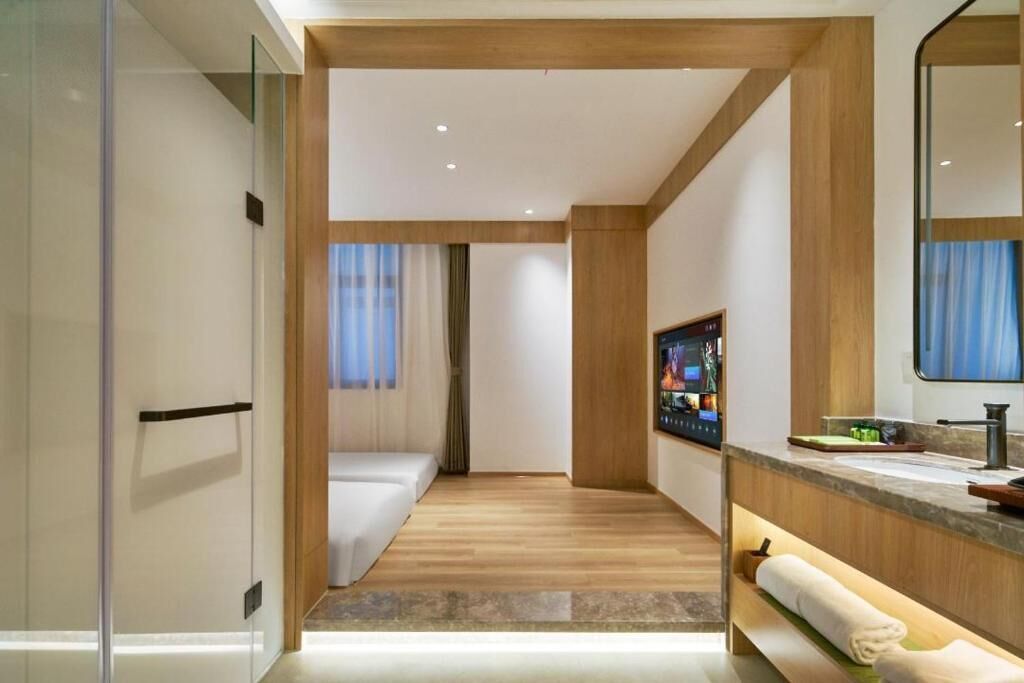 Una habitación de hotel moderna con suelo de madera, un espejo grande, un televisor de pantalla plana y un baño con cabina de ducha de vidrio.