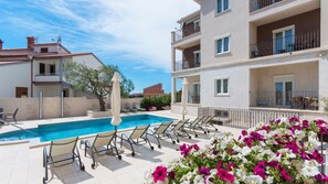 A poolside area with lounge chairs, a pool, and a building with balconies.