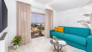 A modern living room with a teal sofa, a flat-screen TV, a small table, and a view of a coastal area.