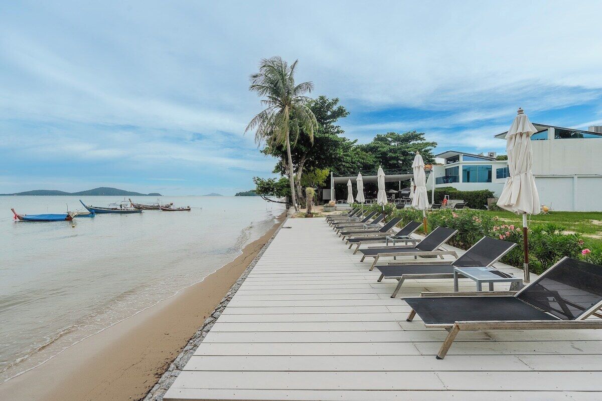 A beachside hotel with a wooden walkway leading to sun loungers and umbrellas, boats in the water, and a clear view of the sea.